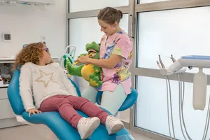 La praticienne explique le brossage à une petite fille au fauteuil avec une peluche crocodile